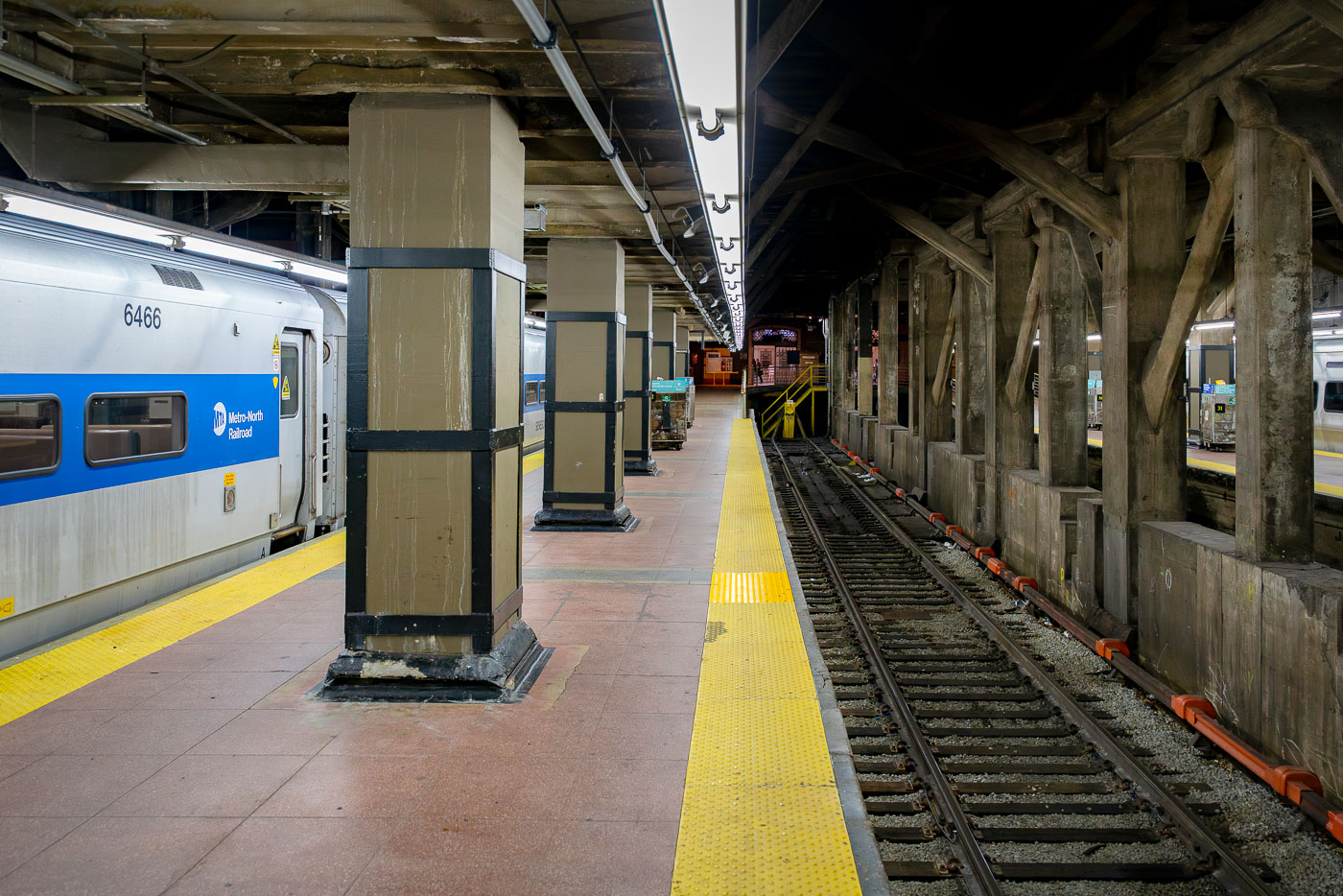 A Metro-North Railroad train waits at a platform inside Grand Central Terminal in Manhattan. The concrete pillars and exposed beams of the underground station frame the tracks, where the yellow warning strip lines the edge of the platform.