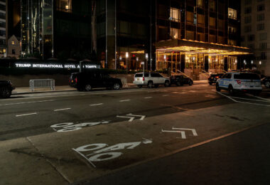 The entrance of Trump International Hotel and Tower in Manhattan is seen at night. Several vehicles, including an NYPD patrol car, are parked outside the building along a marked bike lane.
