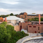 The University of Minnesota’s Southeast Steam Plant sits along the Mississippi River in Minneapolis, supplying heat and power to the campus and nearby medical facilities. Its red-brick structures and tall orange exhaust stacks stand below the university’s historic engineering buildings.