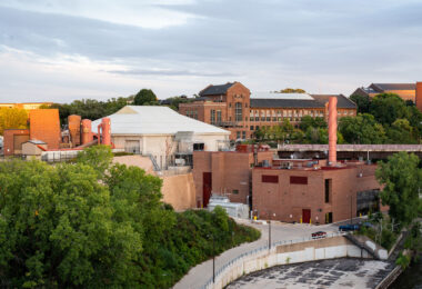 The University of Minnesota’s Southeast Steam Plant sits along the Mississippi River in Minneapolis, supplying heat and power to the campus and nearby medical facilities. Its red-brick structures and tall orange exhaust stacks stand below the university’s historic engineering buildings.