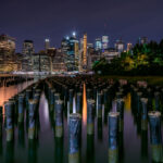 Old wooden pilings rise from the East River with the Manhattan skyline glowing in the background. Viewed from Brooklyn Bridge Park