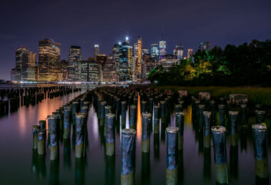 Old wooden pilings rise from the East River with the Manhattan skyline glowing in the background. Viewed from Brooklyn Bridge Park