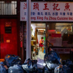 On a dimly lit street in New York’s Chinatown, the glow of a small Fuzhou restaurant spills onto the sidewalk piled high with black garbage bags. Through the narrow doorway of Zheng Xing Fu Zhou Cuisine Inc., a worker in a red apron is seen bent over, quietly preparing food inside the cramped kitchen. The storefront, framed by boxes and handwritten notices, tells the story of a neighborhood balancing survival and tradition, even as the detritus of the day lines the curb outside. The contrast between the bustling work inside and the refuse outside captures a candid snapshot of city life — grit and perseverance side by side.