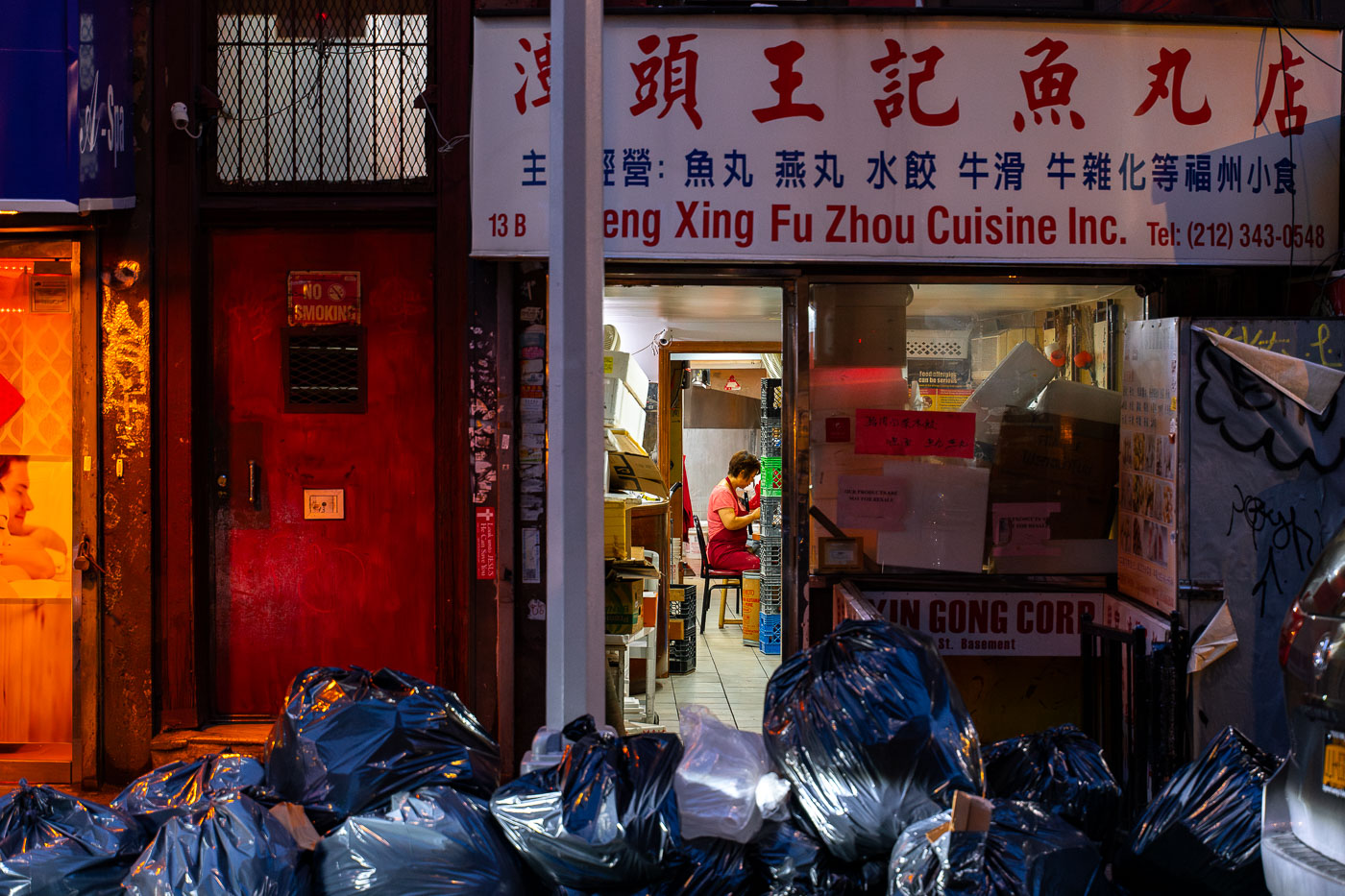 On a dimly lit street in New York’s Chinatown, the glow of a small Fuzhou restaurant spills onto the sidewalk piled high with black garbage bags. Through the narrow doorway of Zheng Xing Fu Zhou Cuisine Inc., a worker in a red apron is seen bent over, quietly preparing food inside the cramped kitchen. The storefront, framed by boxes and handwritten notices, tells the story of a neighborhood balancing survival and tradition, even as the detritus of the day lines the curb outside. The contrast between the bustling work inside and the refuse outside captures a candid snapshot of city life — grit and perseverance side by side.