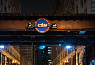 The Chicago Transit Authority (CTA) logo is displayed beneath the steel elevated tracks that define the Loop, the city’s central business district. The structure, part of the “L” system first built in the late 19th century, remains one of Chicago’s most enduring symbols of urban mobility. Overhead girders and riveted beams—many still original to the early 1900s—carry trains above streets lined with early skyscrapers and historic facades. The CTA continues to operate one of the largest and oldest public transit networks in the United States, moving more than a million riders daily through Chicago’s interconnected system of rail and bus lines.