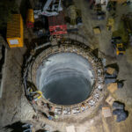 An aerial view captures the excavation shaft for a massive water main tunnel beneath the Mississippi River. The reinforced concrete cylinder serves as the launch point for a tunnel boring machine, visible nearby, which will cut through layers of limestone and shale to create a new conduit linking the city’s water systems. The project represents a critical upgrade to regional infrastructure, designed to improve reliability and resilience while accommodating future demand across Minneapolis and St. Paul.
