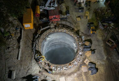 An aerial view captures the excavation shaft for a massive water main tunnel beneath the Mississippi River. The reinforced concrete cylinder serves as the launch point for a tunnel boring machine, visible nearby, which will cut through layers of limestone and shale to create a new conduit linking the city’s water systems. The project represents a critical upgrade to regional infrastructure, designed to improve reliability and resilience while accommodating future demand across Minneapolis and St. Paul.