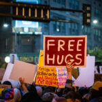 Free Hugs Sign at Trump Protest in Downtown Minneapolis 1 A protester holds a “Free Hugs” sign amid a large demonstration outside the Target Center in Minneapolis on October 10, 2019. The event coincided with Donald Trump’s campaign rally, drawing thousands who gathered in the rain to voice opposition to his administration’s rhetoric and policies. Despite the tense political atmosphere, moments of solidarity and humor emerged throughout the crowd.