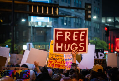 A protester holds a “Free Hugs” sign amid a large demonstration outside the Target Center in Minneapolis on October 10, 2019. The event coincided with Donald Trump’s campaign rally, drawing thousands who gathered in the rain to voice opposition to his administration’s rhetoric and policies. Despite the tense political atmosphere, moments of solidarity and humor emerged throughout the crowd.