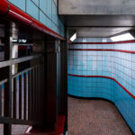 Inside a tiled subway passageway, light blue and red accents curve along the walls, leading commuters toward the platform below. The design is distinctly retro, with polished tiles and fluorescent lighting evoking a mid-20th-century aesthetic found in older metro systems. The turn of the corridor reveals a glimpse of signage and station names, grounding the space in the everyday flow of city life.