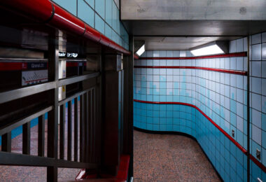 Inside a tiled subway passageway, light blue and red accents curve along the walls, leading commuters toward the platform below. The design is distinctly retro, with polished tiles and fluorescent lighting evoking a mid-20th-century aesthetic found in older metro systems. The turn of the corridor reveals a glimpse of signage and station names, grounding the space in the everyday flow of city life.