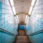 A stairway leads down to the platform at the Chicago Transit Authority’s Grand Station on the Red Line. The tiled corridor—lined with blue ceramic panels and stainless-steel railings—reflects the late-20th-century design style common to CTA subway renovations, emphasizing functionality and durable materials. The bright fluorescent lighting and tiled curvature guide commuters from street level into the subterranean rail network that connects Chicago’s North Side, Loop, and South Side neighborhoods. The subtle skyline motif on the tiles pays homage to the city’s architectural identity while marking one of the Red Line’s busiest downtown access points.