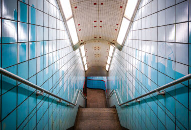 A stairway leads down to the platform at the Chicago Transit Authority’s Grand Station on the Red Line. The tiled corridor—lined with blue ceramic panels and stainless-steel railings—reflects the late-20th-century design style common to CTA subway renovations, emphasizing functionality and durable materials. The bright fluorescent lighting and tiled curvature guide commuters from street level into the subterranean rail network that connects Chicago’s North Side, Loop, and South Side neighborhoods. The subtle skyline motif on the tiles pays homage to the city’s architectural identity while marking one of the Red Line’s busiest downtown access points.