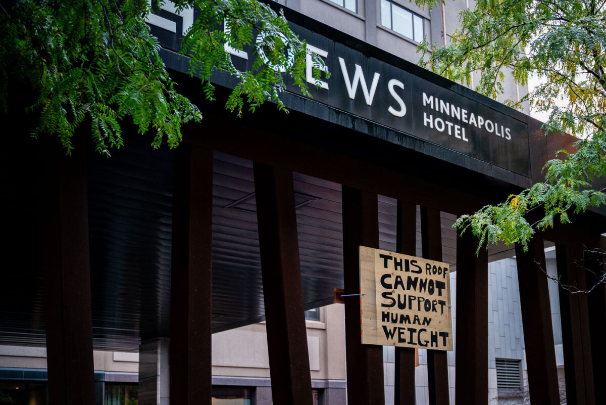 A handmade warning sign reading “This Roof Cannot Support Human Weight” hangs beneath the Loews Minneapolis Hotel canopy on October 10, 2019. The hotel, located adjacent to the Target Center, became a key gathering point during the Trump campaign rally and counter-protests that drew thousands to downtown Minneapolis. City officials placed similar signs throughout the area to prevent demonstrators from climbing elevated structures amid heavy police presence and blocked-off streets.