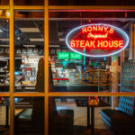 Ronny’s Original Steak House, a longtime fixture of Chicago’s Loop dining scene, glows under its vintage neon sign at night. Founded in 1963, the cafeteria-style steakhouse has served generations of downtown workers, theatergoers, and travelers from its no-frills location near the CTA Red Line and City Hall. Its tufted green booths, mirrored walls, and steady stream of regulars evoke an earlier era of affordable downtown dining—one of the few survivors of mid-century Loop eateries that once defined the city’s commercial core.