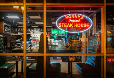Ronny’s Original Steak House, a longtime fixture of Chicago’s Loop dining scene, glows under its vintage neon sign at night. Founded in 1963, the cafeteria-style steakhouse has served generations of downtown workers, theatergoers, and travelers from its no-frills location near the CTA Red Line and City Hall. Its tufted green booths, mirrored walls, and steady stream of regulars evoke an earlier era of affordable downtown dining—one of the few survivors of mid-century Loop eateries that once defined the city’s commercial core.