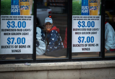 A woman wearing a “Trump 2020” hat smiles from inside a downtown Minneapolis bar near the Target Center on October 10, 2019. The establishment, decorated with promotional posters for discounted beer and wings, became a temporary gathering spot for attendees arriving early to the president’s evening campaign rally. The 2019 visit marked a rare appearance by a sitting U.S. president in Minneapolis and drew both supporters and demonstrators into the city’s entertainment district, where security and anticipation heightened throughout the afternoon.