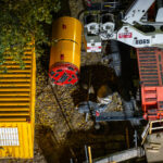 A tunnel boring machine (TBM) rests near the banks of the Mississippi River, staged for the start of a subsurface water main tunnel project. The machine’s circular cutting head and support equipment are visible beside heavy lifting cranes, part of a major infrastructure effort to modernize the city’s underground water distribution system