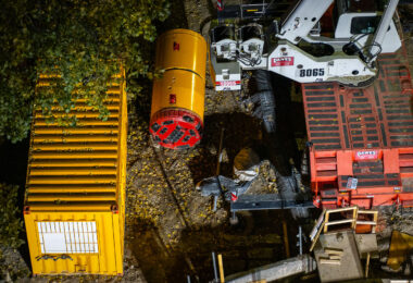 A tunnel boring machine (TBM) rests near the banks of the Mississippi River, staged for the start of a subsurface water main tunnel project. The machine’s circular cutting head and support equipment are visible beside heavy lifting cranes, part of a major infrastructure effort to modernize the city’s underground water distribution system