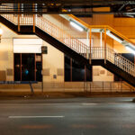 An illuminated stairway leads up to one of Chicago’s iconic “L” (elevated) train platforms in the Loop at night. The steel structure, marked with the system’s signature riveted beams, is part of the century-old elevated rail network that defines the city’s core transit identity. Below, a “Police Line – Do Not Cross” barricade and the quiet, empty street evoke a rare moment of stillness in an area usually dominated by the hum of trains and late-night traffic. This view highlights the utilitarian geometry and enduring presence of the Chicago Transit Authority’s elevated system, still serving as the backbone of the city’s rapid transit since the late 19th century.