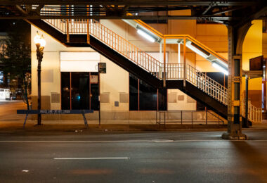 An illuminated stairway leads up to one of Chicago’s iconic “L” (elevated) train platforms in the Loop at night. The steel structure, marked with the system’s signature riveted beams, is part of the century-old elevated rail network that defines the city’s core transit identity. Below, a “Police Line – Do Not Cross” barricade and the quiet, empty street evoke a rare moment of stillness in an area usually dominated by the hum of trains and late-night traffic. This view highlights the utilitarian geometry and enduring presence of the Chicago Transit Authority’s elevated system, still serving as the backbone of the city’s rapid transit since the late 19th century.