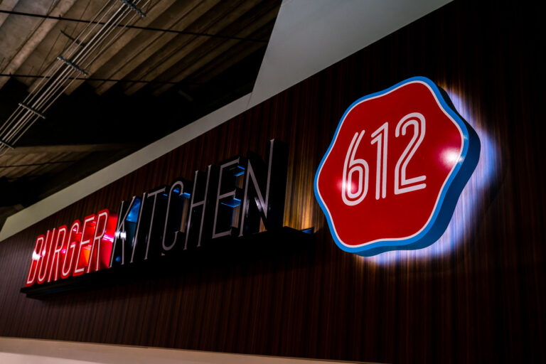 612 Burger Kitchen Sign at U.S. Bank Stadium 1 An illuminated “612 Burger Kitchen” sign inside U.S. Bank Stadium in Minneapolis, named after the city’s area code. The bold red-and-black typography and retro badge design reflect a blend of local identity and modern stadium branding. Located among the venue’s food and beverage concessions, 612 Burger Kitchen is part of the stadium’s effort to feature Minneapolis-based culinary offerings for fans attending Minnesota Vikings games and large-scale events.