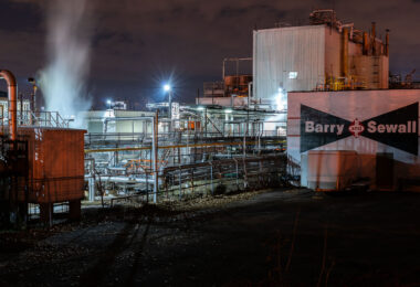 Steam rises from the Barry & Sewall Chemical Company complex in Northeast Minneapolis, a facility known for producing adhesives, coatings, and specialty chemicals for over a century. Illuminated by floodlights and framed by dense pipe networks, the site reflects the city’s enduring industrial infrastructure along the rail and river corridors. The hand-painted logo on the wall remains a remnant of mid-20th-century branding still visible across many older manufacturing sites in the Twin Cities.