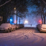 A lone cyclist rides through a snow-covered street in Minneapolis on November 26, 2019, as a major winter storm blankets the city. Streetlights and passing car headlights glow against the falling snow, illuminating parked cars buried in accumulation. The quiet, blue-tinged scene reflects the city’s resilience and rhythm during early-season blizzards that frequently test commuters across the Twin Cities.