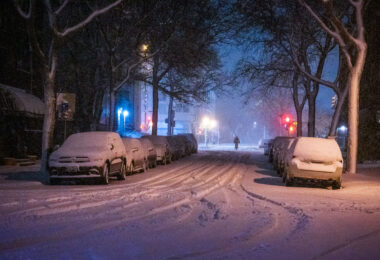 A lone cyclist rides through a snow-covered street in Minneapolis on November 26, 2019, as a major winter storm blankets the city. Streetlights and passing car headlights glow against the falling snow, illuminating parked cars buried in accumulation. The quiet, blue-tinged scene reflects the city’s resilience and rhythm during early-season blizzards that frequently test commuters across the Twin Cities.