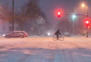 A cyclist crosses Lyndale Avenue South during the heavy November 26, 2019 snowstorm in Minneapolis. Despite low visibility and slick streets, a few commuters still braved the conditions, illuminated by red traffic lights and snow-diffused streetlamps. The image captures the perseverance of winter cycling culture in the Twin Cities, where even severe weather rarely stops movement through the urban grid.