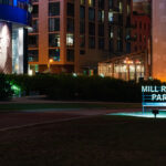 The illuminated sign for Mill Ruins Park glows at night along the Minneapolis riverfront, where historic flour mill foundations meet modern development. Behind it, a portrait mural and new residential buildings rise near the preserved remnants of the city’s milling heyday.