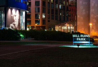 The illuminated sign for Mill Ruins Park glows at night along the Minneapolis riverfront, where historic flour mill foundations meet modern development. Behind it, a portrait mural and new residential buildings rise near the preserved remnants of the city’s milling heyday.