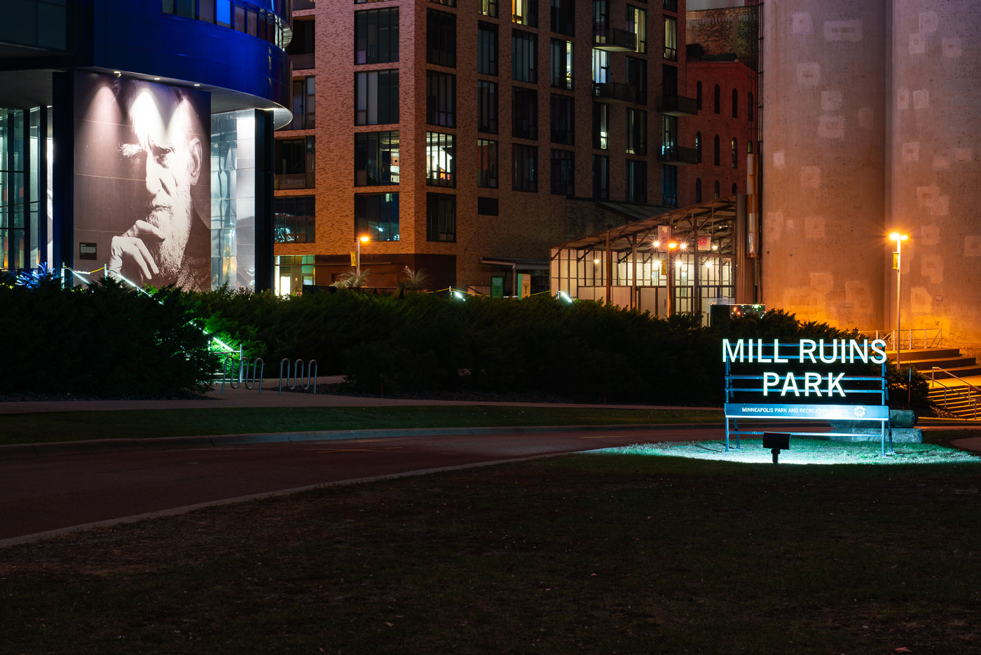 The illuminated sign for Mill Ruins Park glows at night along the Minneapolis riverfront, where historic flour mill foundations meet modern development. Behind it, a portrait mural and new residential buildings rise near the preserved remnants of the city’s milling heyday.