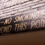 A weathered metal sign mounted on a brick wall warns “NO SMOKING BEYOND THIS POINT,” its paint cracked and dulled by decades of exposure. Signs like this were once common in milling and industrial facilities across Minneapolis, where airborne dust from grain or manufacturing posed a significant fire risk. The hand-lettered typography and aged surface reflect a bygone era of factory safety culture, preserving a small but telling fragment of the city’s industrial heritage.