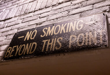 A weathered metal sign mounted on a brick wall warns “NO SMOKING BEYOND THIS POINT,” its paint cracked and dulled by decades of exposure. Signs like this were once common in milling and industrial facilities across Minneapolis, where airborne dust from grain or manufacturing posed a significant fire risk. The hand-lettered typography and aged surface reflect a bygone era of factory safety culture, preserving a small but telling fragment of the city’s industrial heritage.