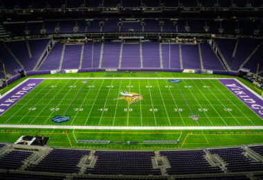 The interior of U.S. Bank Stadium in Minneapolis, home of the NFL’s Minnesota Vikings, shown from a high vantage point with an unobstructed view of the field. Completed in 2016 on the former site of the Metrodome, the $1.1 billion stadium features a fixed, translucent ETFE roof and steeply angled seating designed to evoke Nordic longhouses. The field displays the Vikings’ horned helmet logo at midfield, surrounded by vibrant purple end zones and the signature “SKOL” rally cry—a modern landmark of downtown Minneapolis and a hub for major sporting and entertainment events.