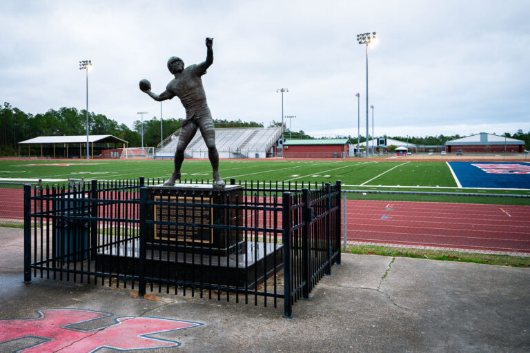 Brett Favre Statue in Kiln Mississippi 1 In Kiln, Mississippi, the legacy of Brett Favre looms large. Outside Hancock High School’s football stadium, a life-sized statue of the quarterback stands mid-throw, immortalizing the local athlete who went on to become one of the NFL’s most prolific passers. Set against the backdrop of the school’s red track and green turf, the monument reflects both community pride and the enduring place of football in Southern culture. For locals, it serves as a reminder that even from a small Gulf Coast town, greatness can reach the national stage.