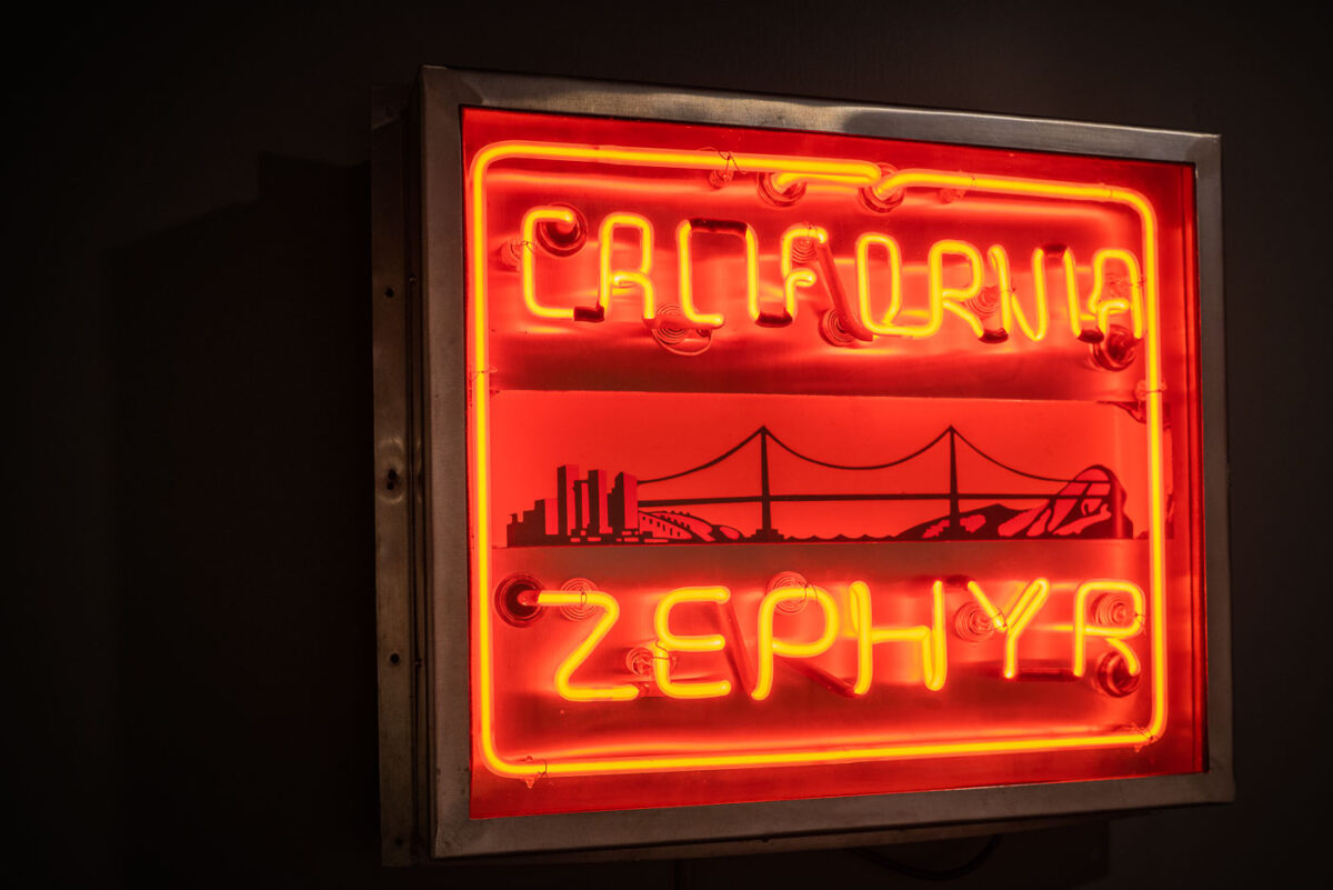 This neon sign once promoted the California Zephyr, one of the most celebrated passenger trains of the postwar era. First introduced in 1949, the train linked Chicago to the San Francisco Bay Area, offering panoramic views of the Rockies and the Sierra Nevada. The glowing red lettering, paired with imagery of a bridge, skyline, and streamlined locomotive, reflects the optimism of mid-20th-century American rail travel and the romance of crossing the continent in comfort and style.