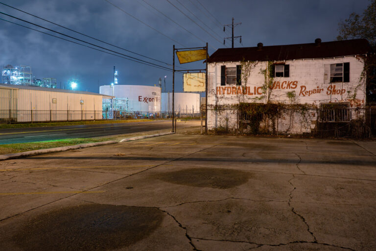 ExxonMobil Refinery and Hydraulic Shop, Baton Rouge 1 An abandoned hydraulic jack repair shop sits across from the massive ExxonMobil refinery complex in Baton Rouge, Louisiana. The plant, one of the largest oil refineries in the United States, has operated since 1909 and remains a central fixture of the city’s industrial identity. The decaying storefront in the foreground contrasts sharply with the illuminated refinery tanks and piping behind it — a visual divide between small-scale industry of the past and the enduring scale of the petrochemical operations that define the region’s economy.