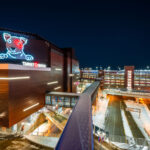 The Target Center’s neon Bullseye dog glows above 1st Avenue North, marking one of Minneapolis’ most recognizable downtown landmarks. Opened in 1990 and renovated in 2017 with a modern metal facade, the arena is home to the Minnesota Timberwolves and a hub for concerts and civic events. In the foreground, the enclosed skyway leads to Ramp A — part of the city’s expansive elevated walkway network that connects much of downtown. Snow-lined streets and the layered lighting of the ramps emphasize Minneapolis’ engineered adaptation to both density and winter.