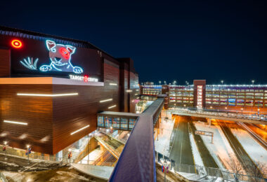 The Target Center’s neon Bullseye dog glows above 1st Avenue North, marking one of Minneapolis’ most recognizable downtown landmarks. Opened in 1990 and renovated in 2017 with a modern metal facade, the arena is home to the Minnesota Timberwolves and a hub for concerts and civic events. In the foreground, the enclosed skyway leads to Ramp A — part of the city’s expansive elevated walkway network that connects much of downtown. Snow-lined streets and the layered lighting of the ramps emphasize Minneapolis’ engineered adaptation to both density and winter.