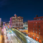 The illuminated Target Field sign overlooks the Warehouse District in downtown Minneapolis, where the city’s historic brick warehouses meet modern transit and redevelopment. Below, the Metro Green and Blue Line trains stop at Target Field Station, a multimodal hub linking light rail, commuter rail, and bike paths. The brightly lit Ford Center and preserved brick structures along 5th Street North illustrate the district’s mix of 20th-century industrial architecture and contemporary urban life, framed against the cold clarity of a Minnesota winter night.