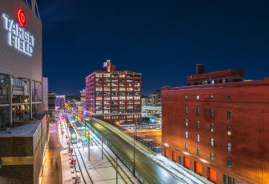 The illuminated Target Field sign overlooks the Warehouse District in downtown Minneapolis, where the city’s historic brick warehouses meet modern transit and redevelopment. Below, the Metro Green and Blue Line trains stop at Target Field Station, a multimodal hub linking light rail, commuter rail, and bike paths. The brightly lit Ford Center and preserved brick structures along 5th Street North illustrate the district’s mix of 20th-century industrial architecture and contemporary urban life, framed against the cold clarity of a Minnesota winter night.