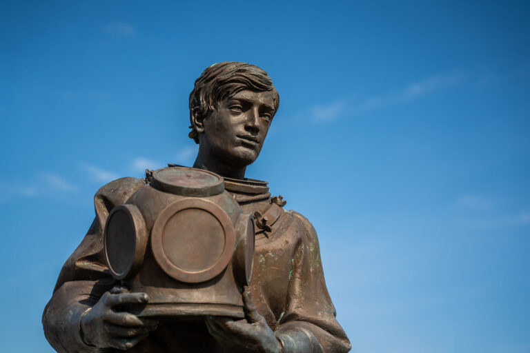Tarpon Springs Sponge Diver Memorial Statue 2 The Sponge Diver Memorial in Tarpon Springs, Florida, honors the Greek immigrants who established the town’s sponge diving industry in the early 1900s. The bronze statue depicts a diver holding a traditional brass helmet, symbolizing the skill and danger of the underwater trade. Located near the historic Sponge Docks, it serves as a tribute to the workers who helped shape the community’s maritime heritage.