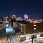On a cold winter night in Minneapolis, the Salvation Army’s brick building stands in the foreground, a reminder of the nonprofit’s long presence serving those in need near the city’s downtown core. Beyond it, steam rises from the nearby power plant while the illuminated skyline glows with high-rise towers, blending industrial grit with modern glass. The juxtaposition of snowy side streets, mid-century warehouses, and gleaming skyscrapers reflects the city’s layered history — from its working-class roots to its contemporary role as a regional hub of commerce and culture.