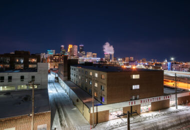 On a cold winter night in Minneapolis, the Salvation Army’s brick building stands in the foreground, a reminder of the nonprofit’s long presence serving those in need near the city’s downtown core. Beyond it, steam rises from the nearby power plant while the illuminated skyline glows with high-rise towers, blending industrial grit with modern glass. The juxtaposition of snowy side streets, mid-century warehouses, and gleaming skyscrapers reflects the city’s layered history — from its working-class roots to its contemporary role as a regional hub of commerce and culture.