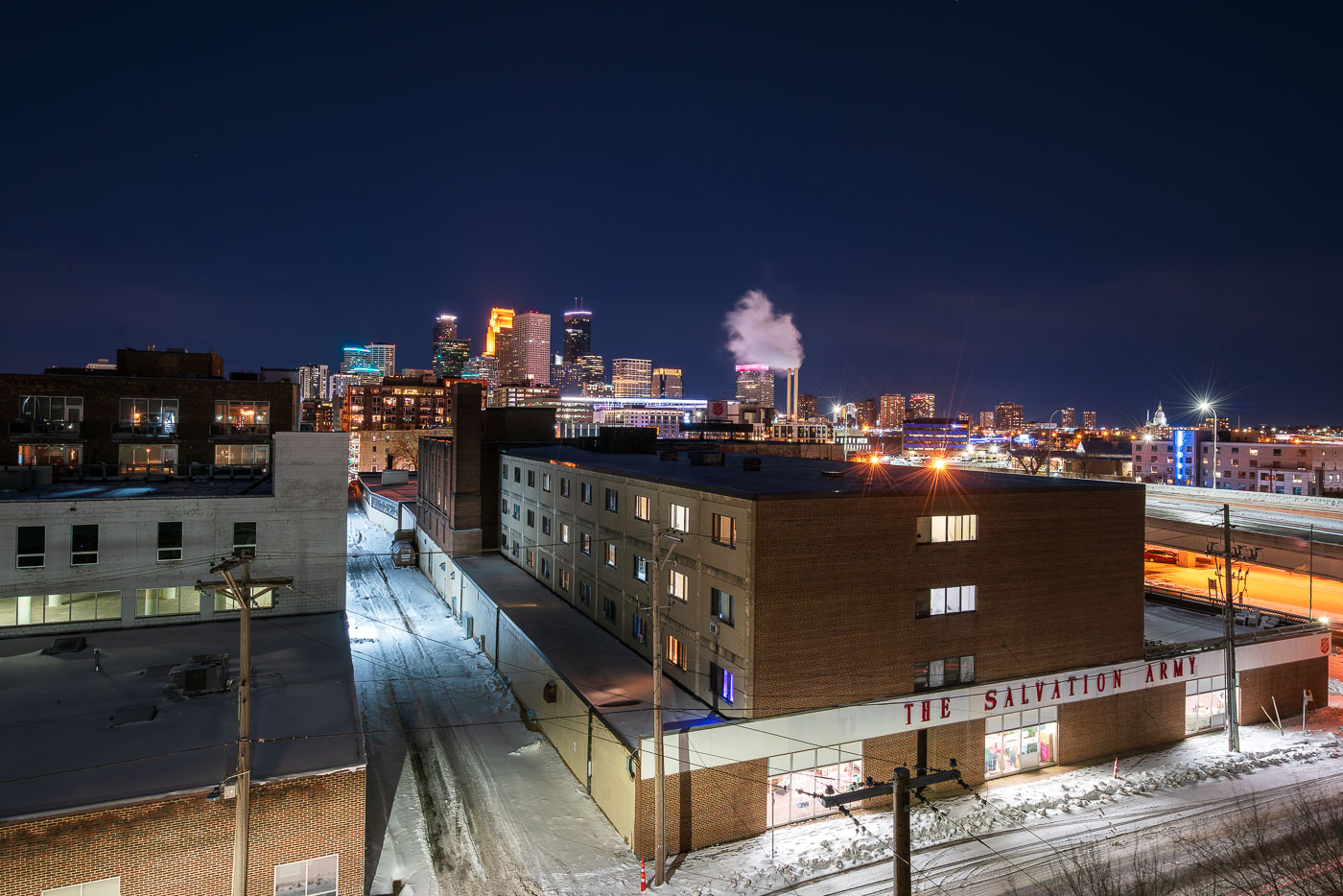 On a cold winter night in Minneapolis, the Salvation Army’s brick building stands in the foreground, a reminder of the nonprofit’s long presence serving those in need near the city’s downtown core. Beyond it, steam rises from the nearby power plant while the illuminated skyline glows with high-rise towers, blending industrial grit with modern glass. The juxtaposition of snowy side streets, mid-century warehouses, and gleaming skyscrapers reflects the city’s layered history — from its working-class roots to its contemporary role as a regional hub of commerce and culture.