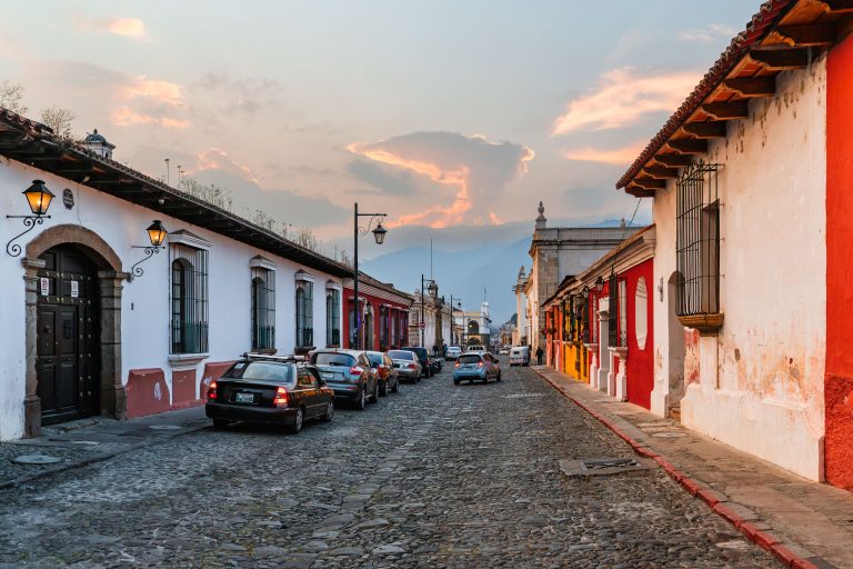Cobblestone Street at Sunset, Antigua Guatemala 4 Colonial street with white and red buildings, parked cars, and church towers at sunset in a Latin American town.