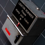 Location Reminder ticket stamping 3 “Location Reminder” ticket stamping machine, once used in transit stations to help passengers remember where they parked or to mark their travel tickets with time and place. The metal box features a lever-operated stamping mechanism, a faded instructional label, and worn edges that show decades of use. Devices like this were common in train station parking facilities and older public transit hubs before digital systems replaced manual timestamping. The angled mounting on tiled wall, along with the utilitarian industrial design, reflects mid-20th-century station infrastructure, where functional machines like this played a part in daily commuter routines.