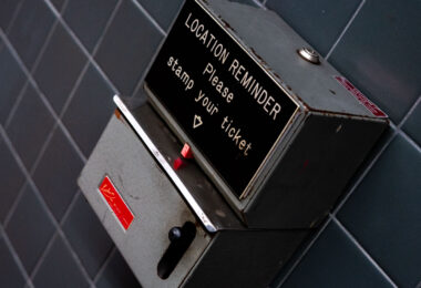 “Location Reminder” ticket stamping machine, once used in transit stations to help passengers remember where they parked or to mark their travel tickets with time and place. The metal box features a lever-operated stamping mechanism, a faded instructional label, and worn edges that show decades of use. Devices like this were common in train station parking facilities and older public transit hubs before digital systems replaced manual timestamping. The angled mounting on tiled wall, along with the utilitarian industrial design, reflects mid-20th-century station infrastructure, where functional machines like this played a part in daily commuter routines.